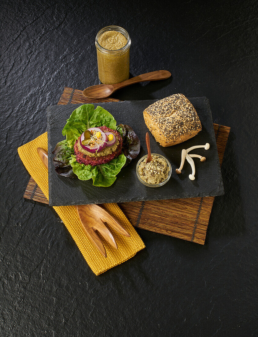 Von oben köstlicher Burger, bestehend aus frischem Salat, Steak, Essiggurken und Zwiebeln mit Soße und Pilzen, auf einem schwarzen Teller neben einem Senfglas