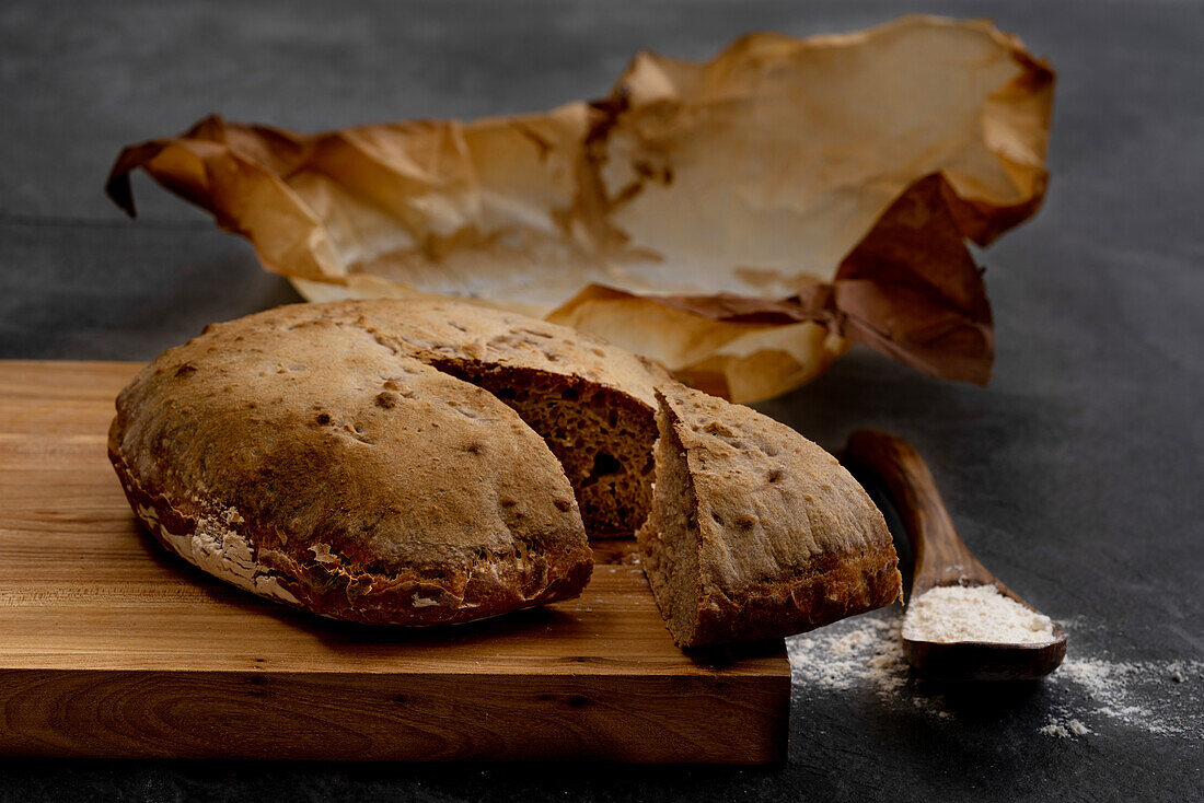 Komposition mit frisch gebackenem, rustikalem, rundem Sauerteigbrot auf Pergamentpapier auf einem Holzbrett mit Löffel und Weizenmehl