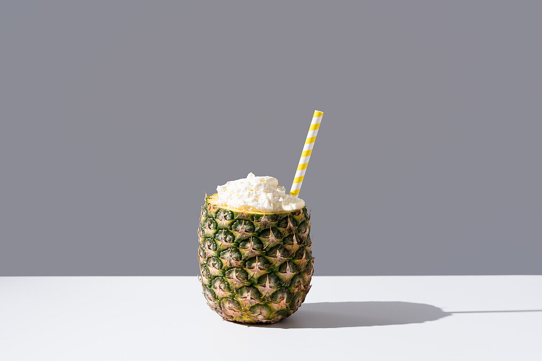 Reife Ananas mit gestreiftem Strohhalm und Sahne auf einem sonnenbeschienenen Tisch, der ein frisches, gesundes Getränk auf grauem Hintergrund symbolisiert