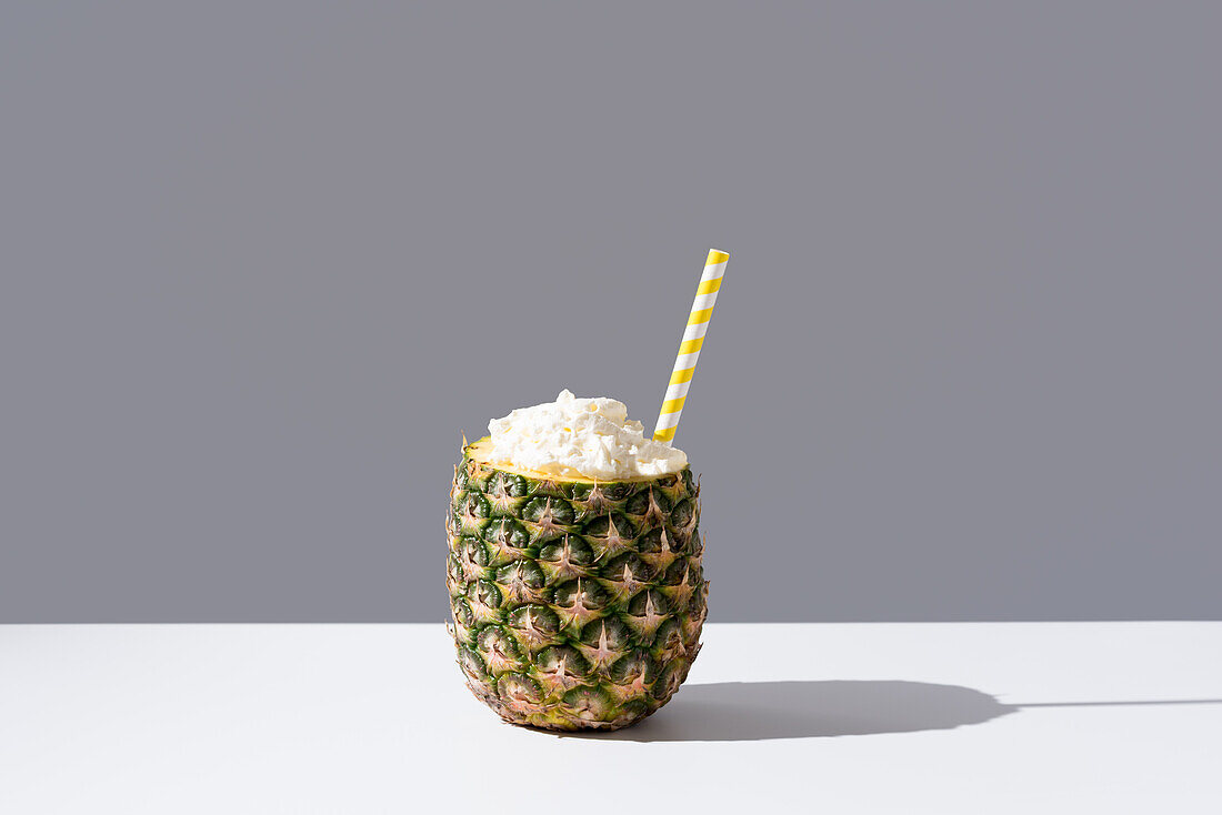 Ripe pineapple with striped straw and cream placed on sunlit table symbolizing fresh healthy drink on gray background