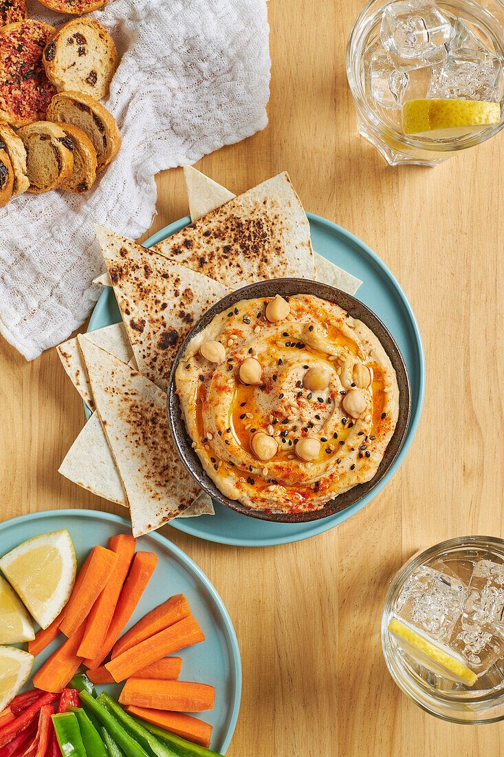 Top view of delicious hummus and tortilla chips served on plate near assorted vegetables and croutons and glasses of cold lemonade on wooden table