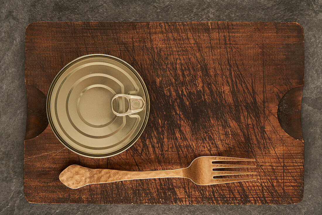 From above scratched chopping board with fork and sealed can with preserved food on rustic lumber table