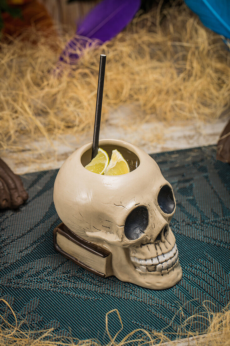 Ceramic polynesian tiki cup skull shaped with straw placed amidst dry grass with wooden fence and colorful feathers on blurred background