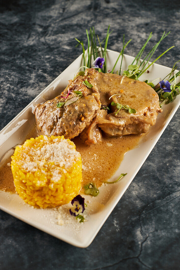 Von oben köstliche Ossobuco Gericht mit Gemüse und Reis-Sauce garniert mit Blumen auf rechteckigen Teller auf Marmortisch während des Mittagessens im Restaurant
