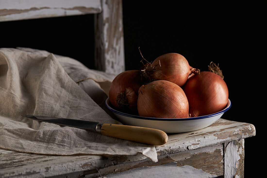 Bowl with whole unpeeled onions placed near linen napkin and knife on shabby wooden chair