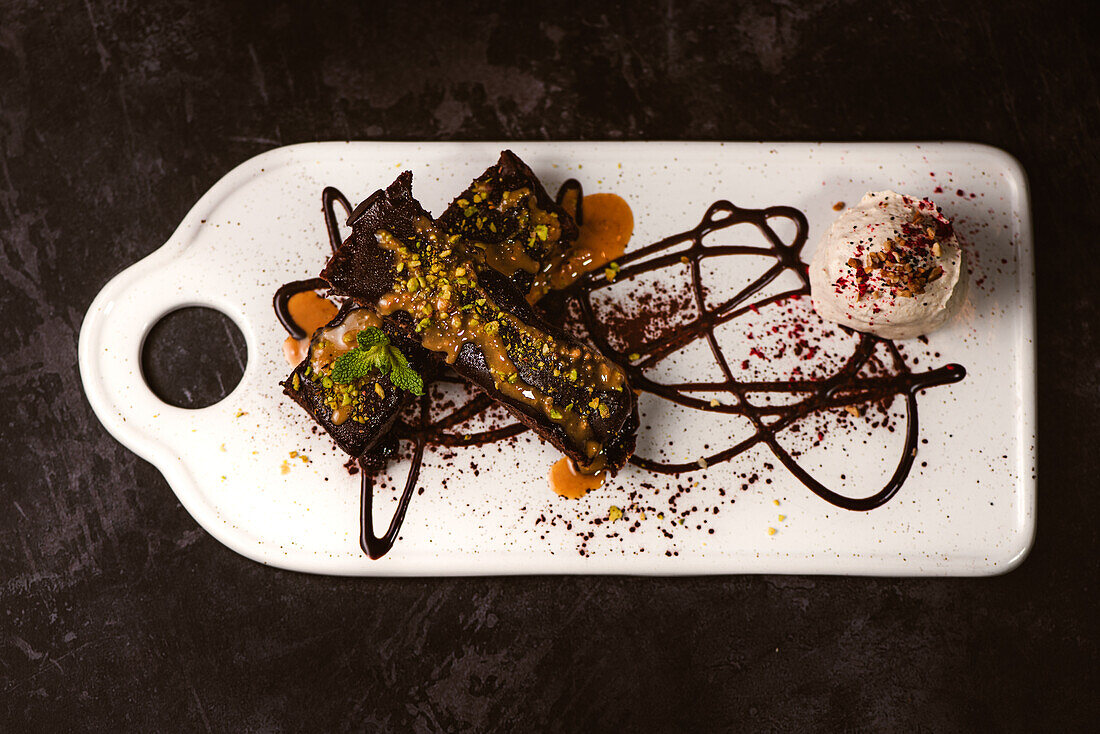 Tasty brownies covered with peanut butter and crunchy crushed pistachios near gelato scoop on plate with chocolate sauce