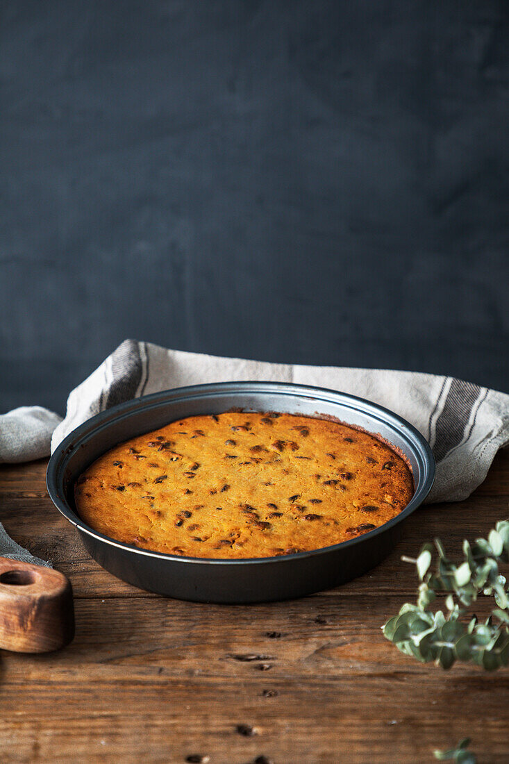 Round tin with freshly baked pumpkin pie placed on rustic lumber table near linen towel and twig against gray background