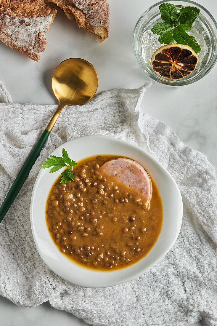 From above of spoon placed near bowl of yummy lentil soup served with slice of sausage and leaf of parsley on marble table and gray napkin