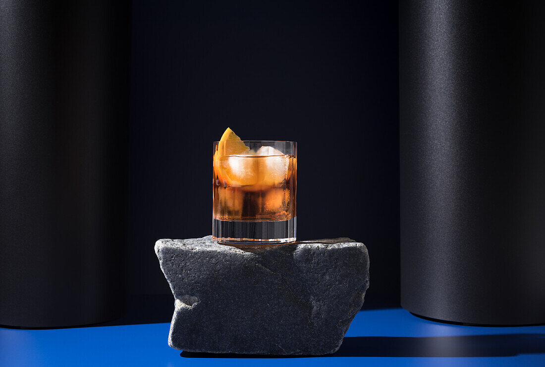 Glass of alcoholic cocktail made with red vermouth served with orange slices on stone in light studio with blue and black wall