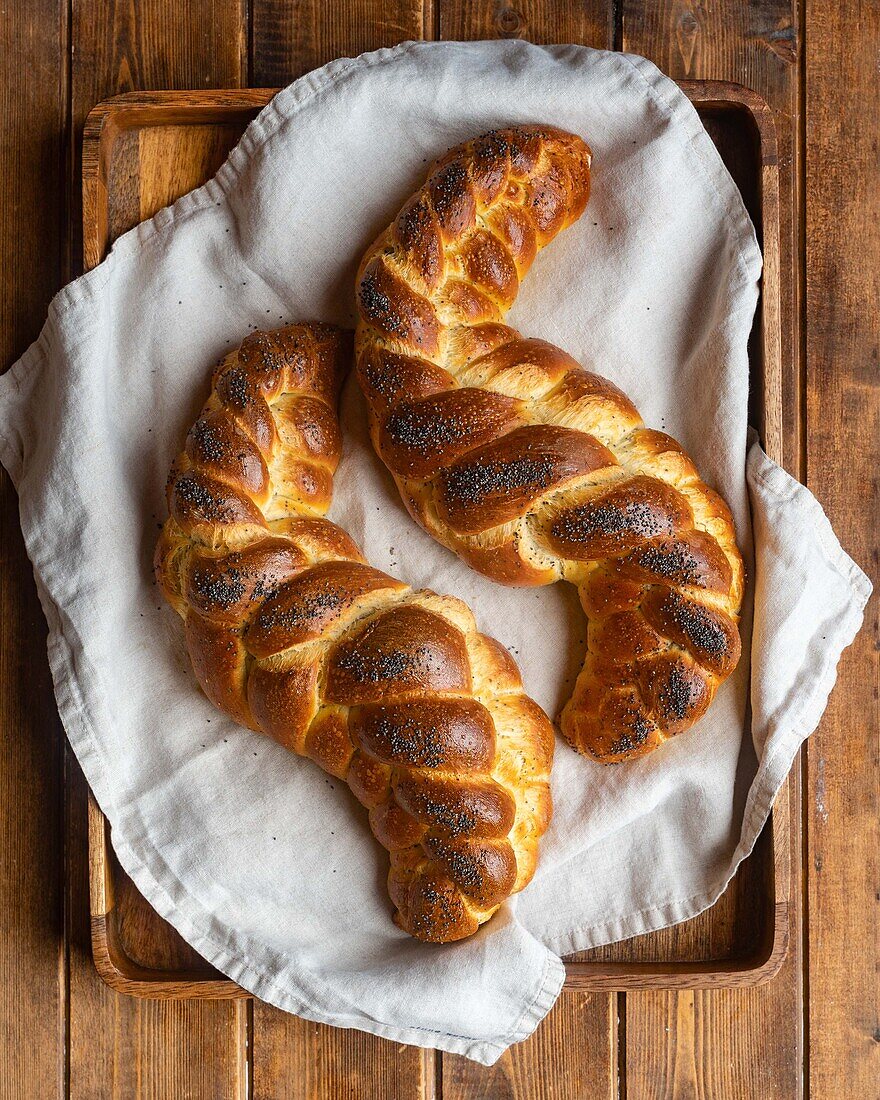 From above of served golden brown bread braids challah with poppy seeds on white napkin in tray