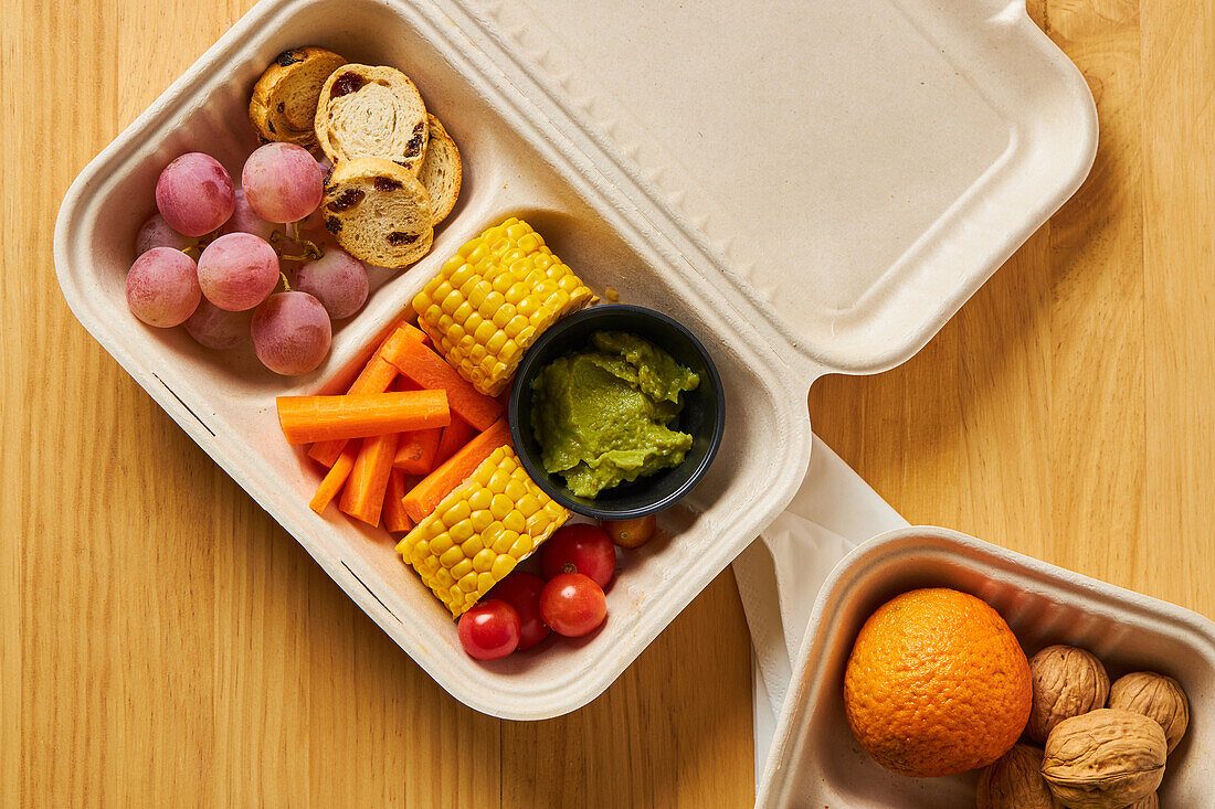 From above of lunch boxes with healthy food including crackers carrot sticks grapes cherry tomatoes with kiwi broccoli walnut and tangerine on yellow background