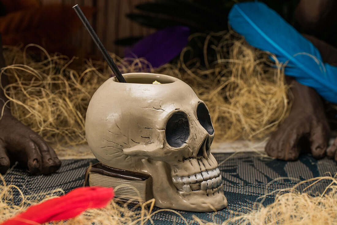 Ceramic polynesian tiki cup skull shaped with straw placed amidst dry grass with wooden fence and colorful feathers on blurred background