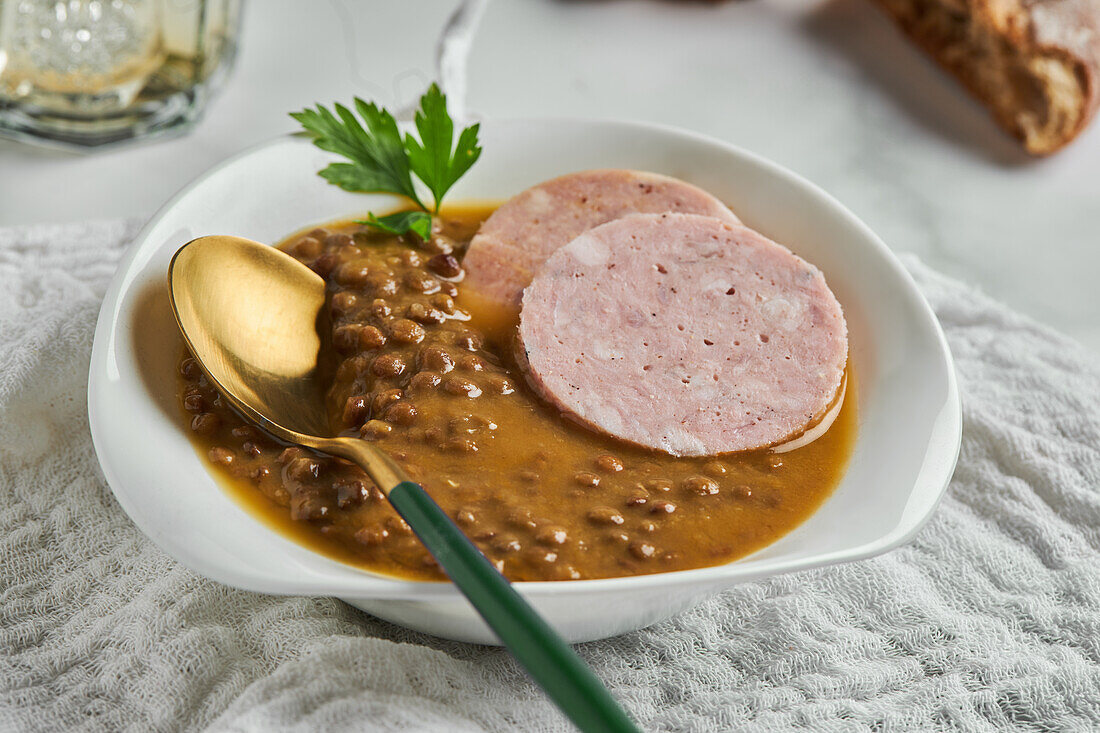 From above spoon placed in bowl of yummy lentil soup served with slices of sausage and leaf of parsley on marble table and gray napkin