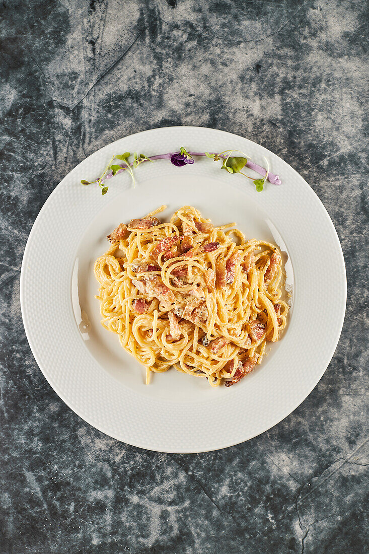 High angle of yummy spaghetti carbonara dish with bacon and parmesan mixed with egg served on plate with sprouts and flower during lunch in Italian restaurant