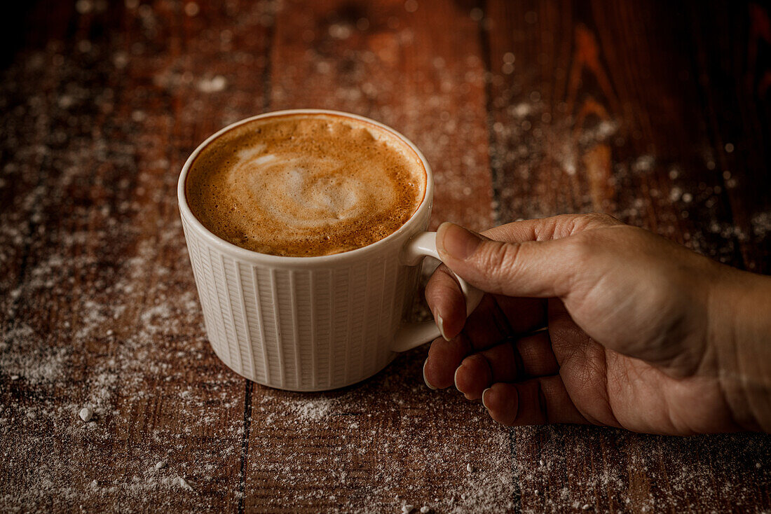 Anonyme Person hält eine Tasse mit aromatischem Kaffee und Schaum auf einem Holztisch
