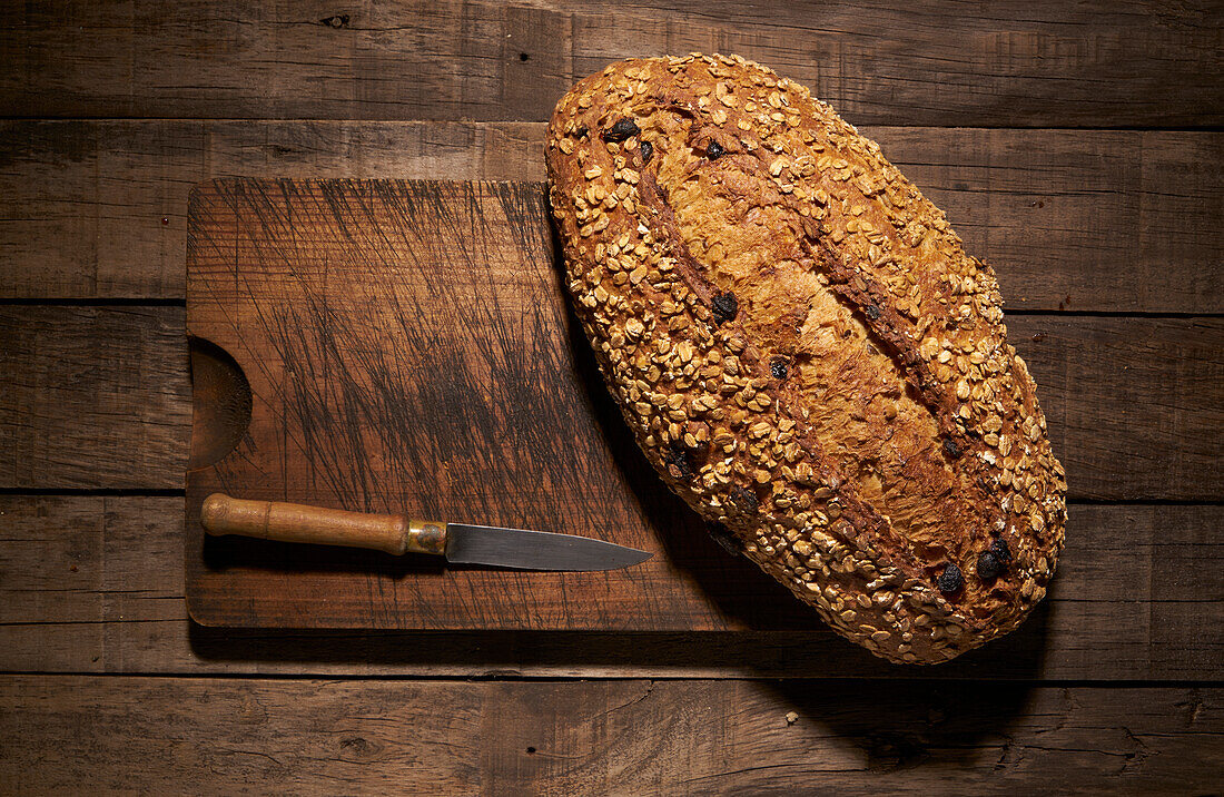 Von oben appetitlich knusprig frisch gebackenen hausgemachten Brotlaib mit Haferflocken und Rosinen mit Messer auf hölzernen Schneidebrett platziert