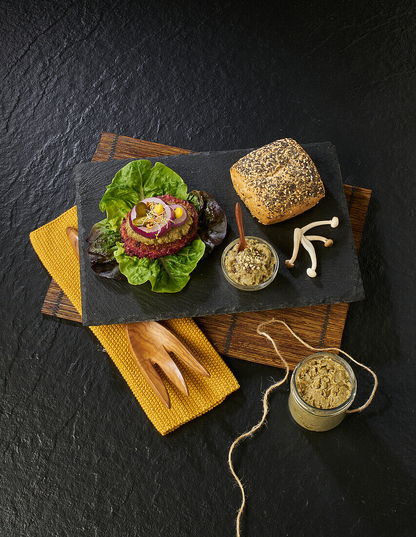 Von oben köstlicher Burger, bestehend aus frischem Salat, Steak, Essiggurken und Zwiebeln mit Soße und Pilzen, auf einem schwarzen Teller neben einem Senfglas