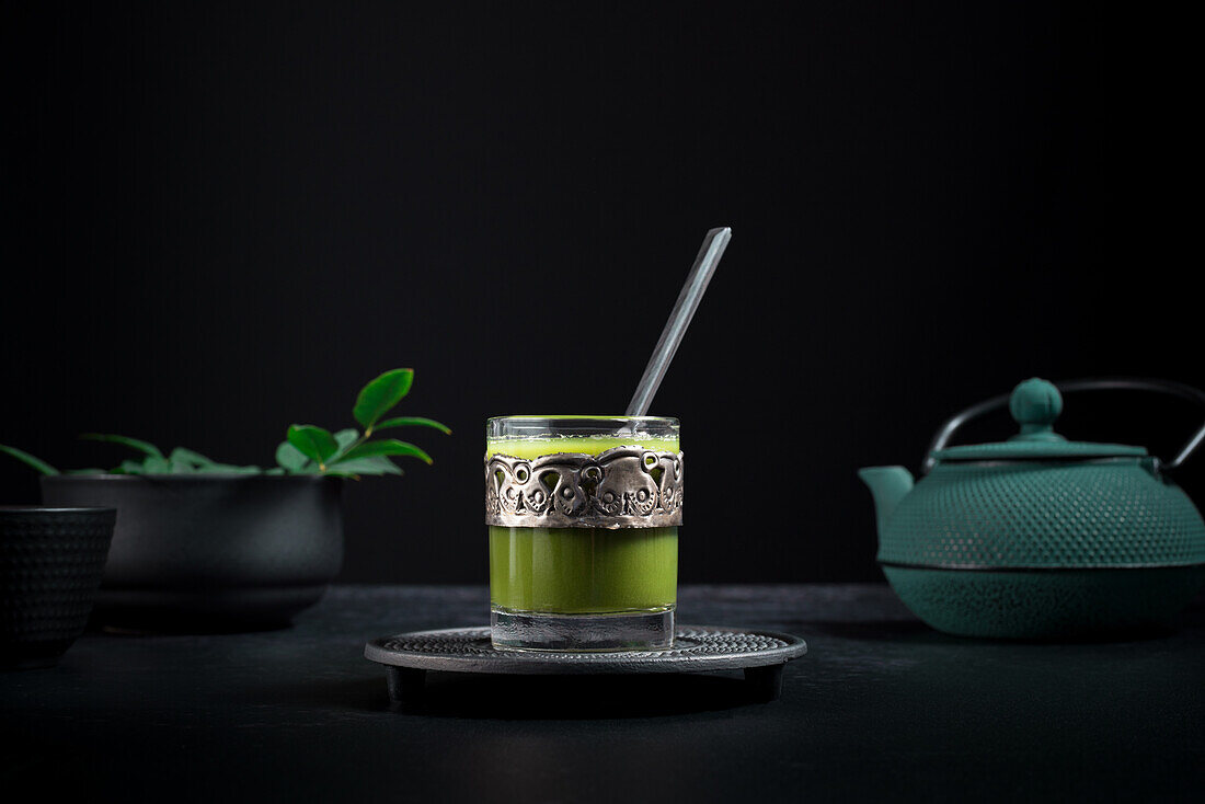 Still life composition with traditional oriental matcha tea served in glass cup with metal ornamental decor on table with ceramic bowls and fresh green leaves against black background