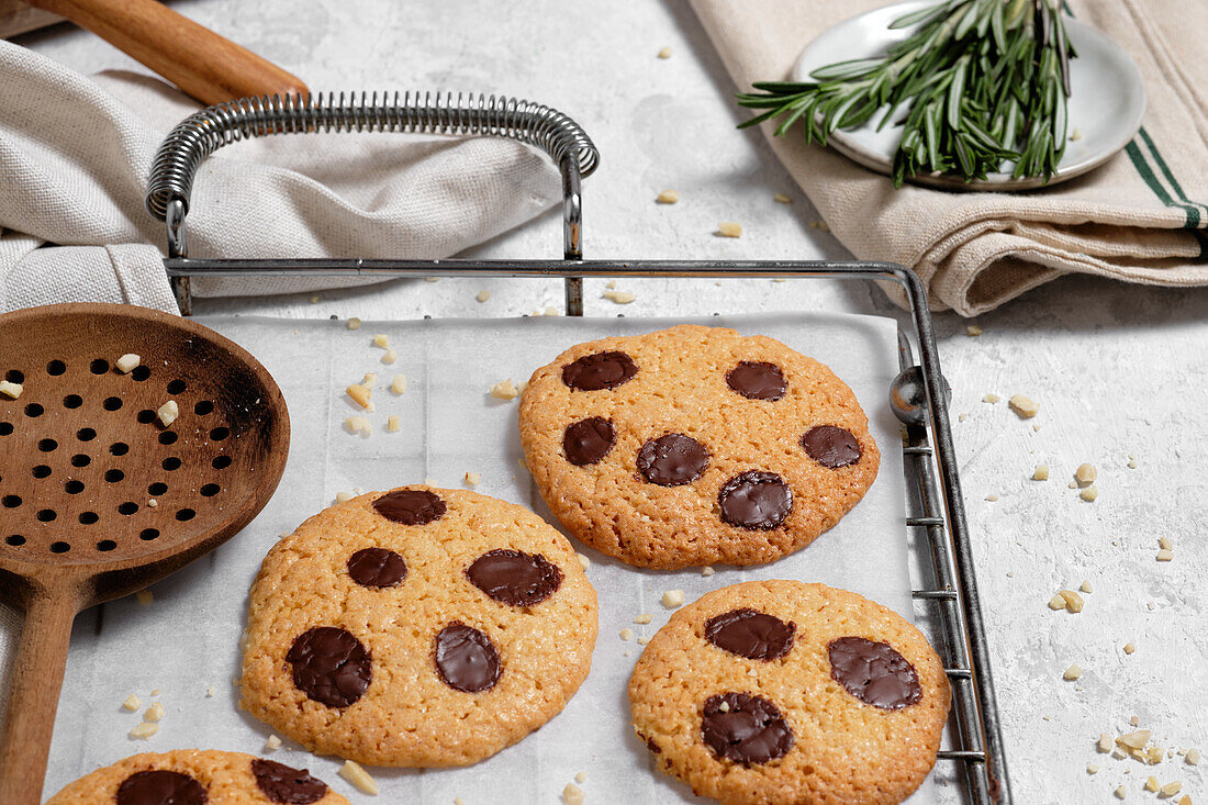 From above of freshly baked sweet cookies with chocolate chips on metal grid placed on table with various kitchen tools and green rosemary branches