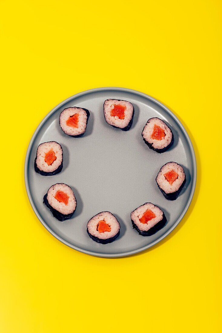 Draufsicht auf eine Portion köstlicher Sushi-Rollen mit Reis und Lachs, eingewickelt in Nori, serviert auf einem grauen Keramikteller auf gelber Oberfläche