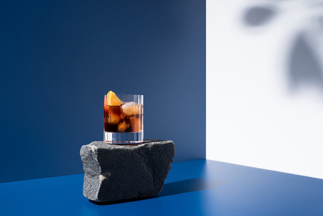 Glass of alcoholic cocktail made with red vermouth served with orange slices on stone in light studio with blue and white wall with shadow