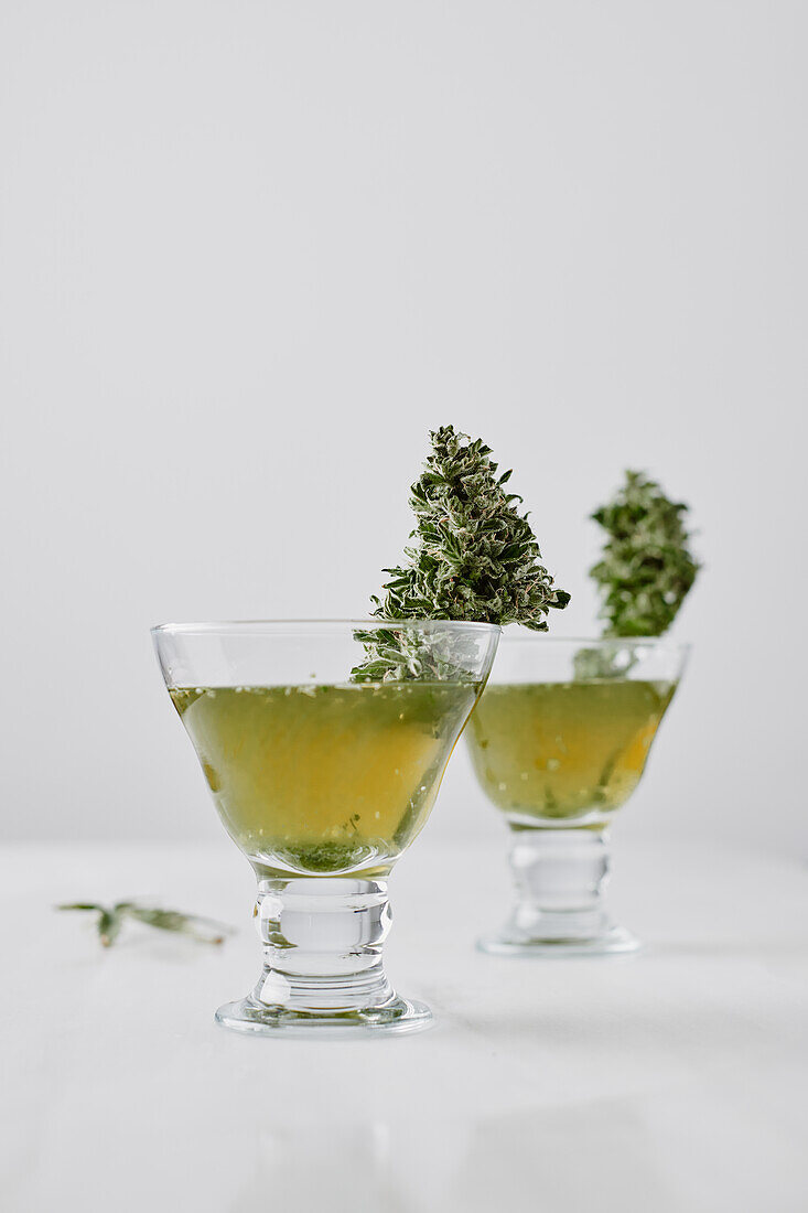 Small glasses with medical marijuana tea and green herbs placed on white surface near glass jar