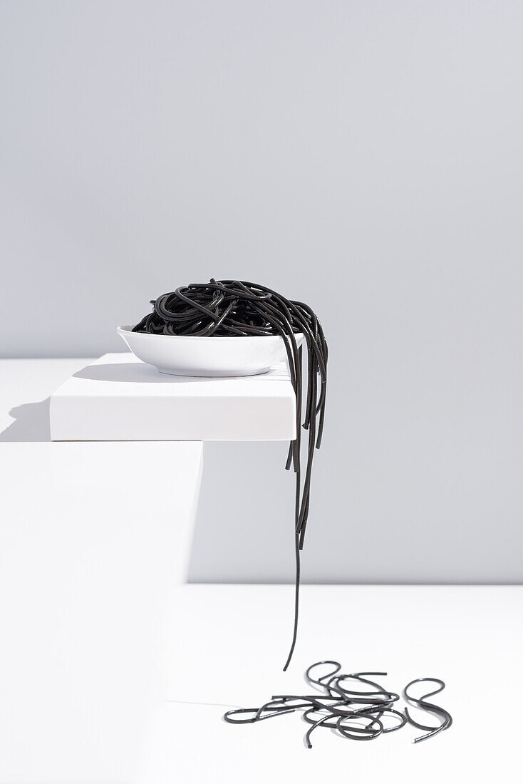 Minimalistisches Atelier mit schwarzen Tintenfischspaghetti, die aus einer vollen Keramikschüssel auf einen weißen Tisch fallen