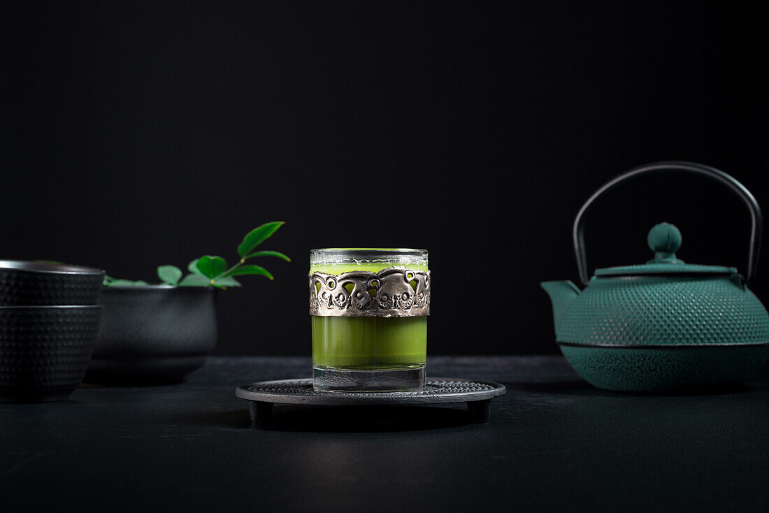 Still life composition with traditional oriental matcha tea served in glass cup with metal ornamental decor on table with ceramic bowls and fresh green leaves against black background