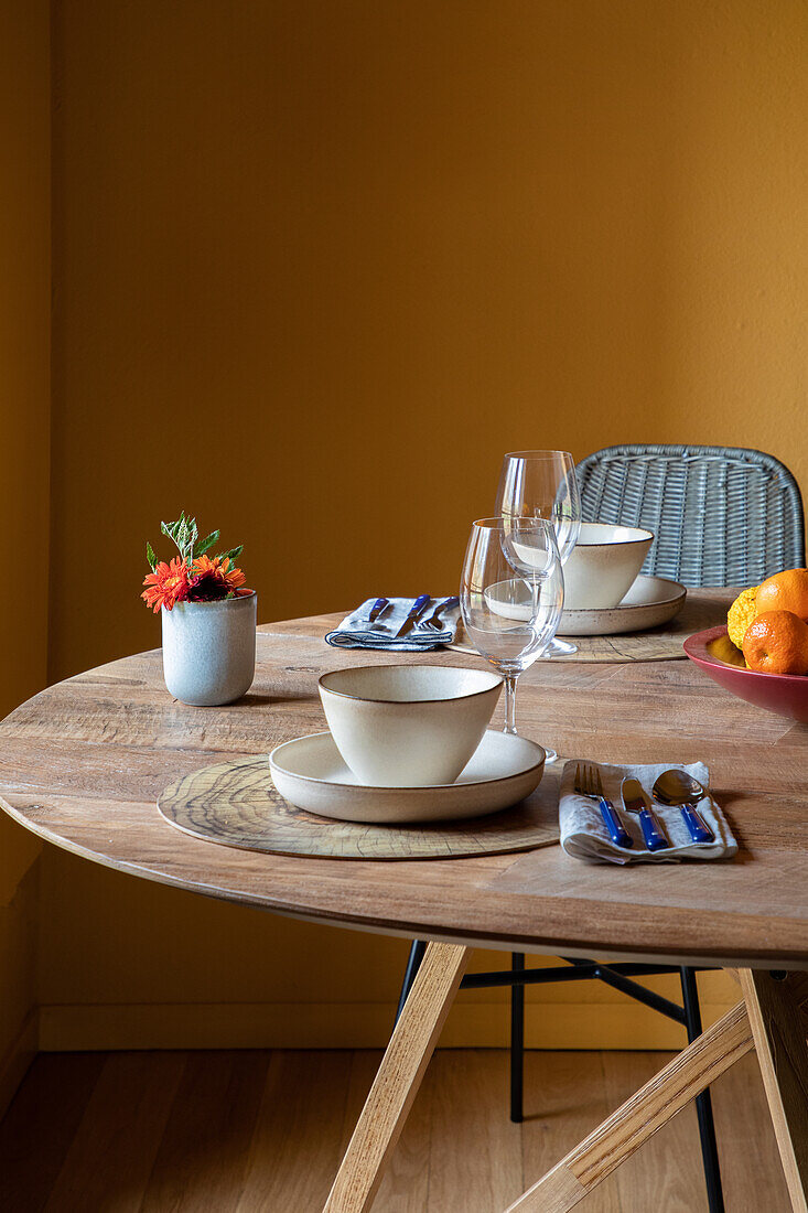 Servierter Tisch mit Keramikschalen auf Tellern mit Besteck auf Serviette neben Weingläsern und Blumen mit Früchten