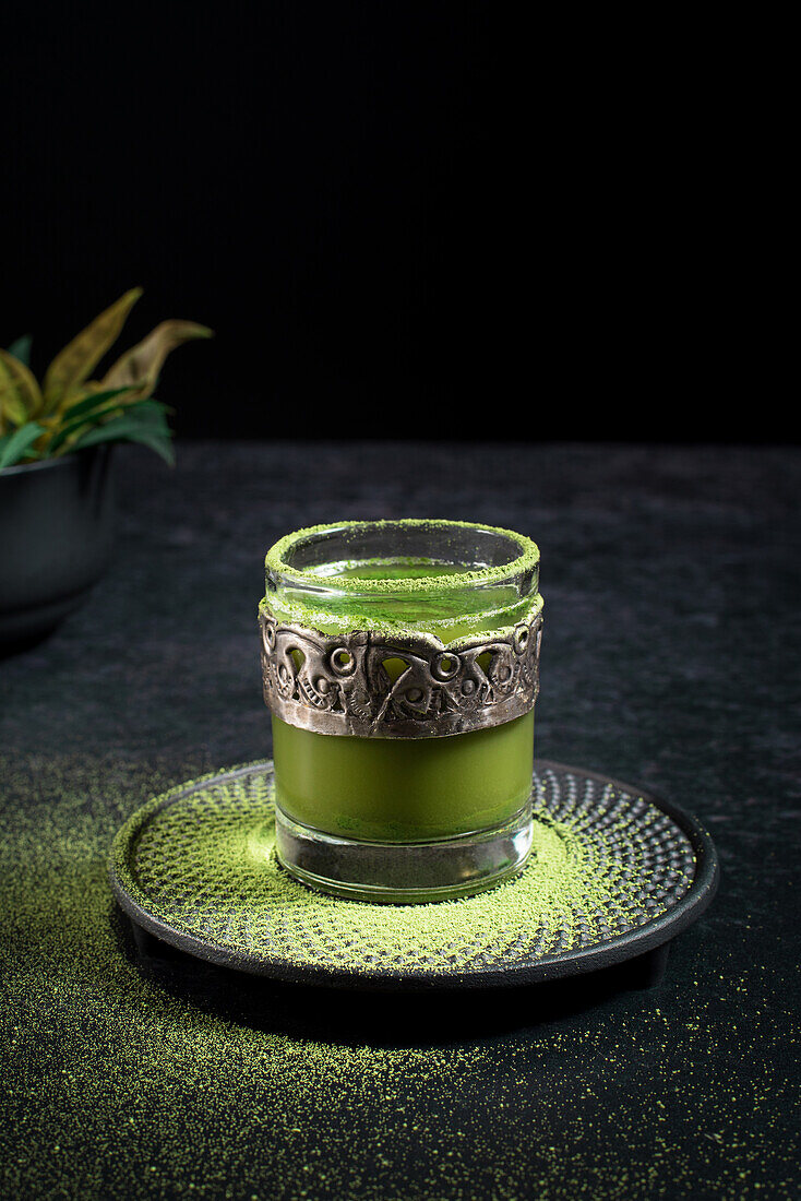 From above of healthy herbal green matcha tea served in glass cup with metal decoration on saucer sprinkled with powder on black table