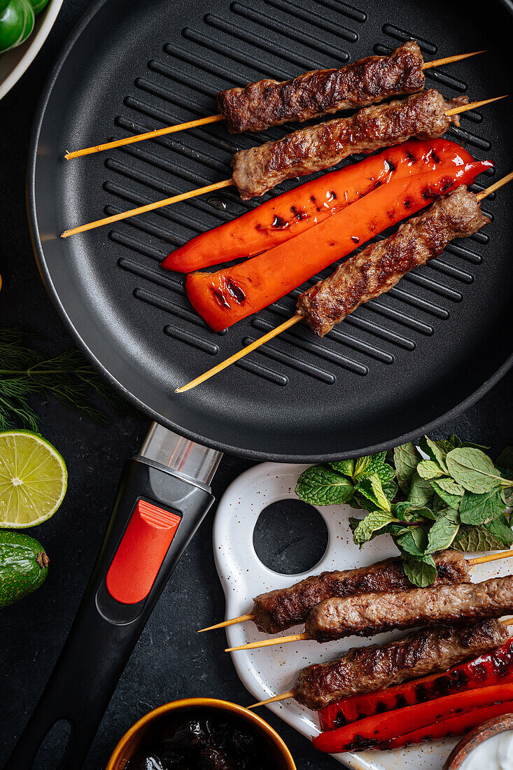 Top view of tasty roasted kebab on skewers and red pepper on grill pan placed on table with assorted vegetables in kitchen