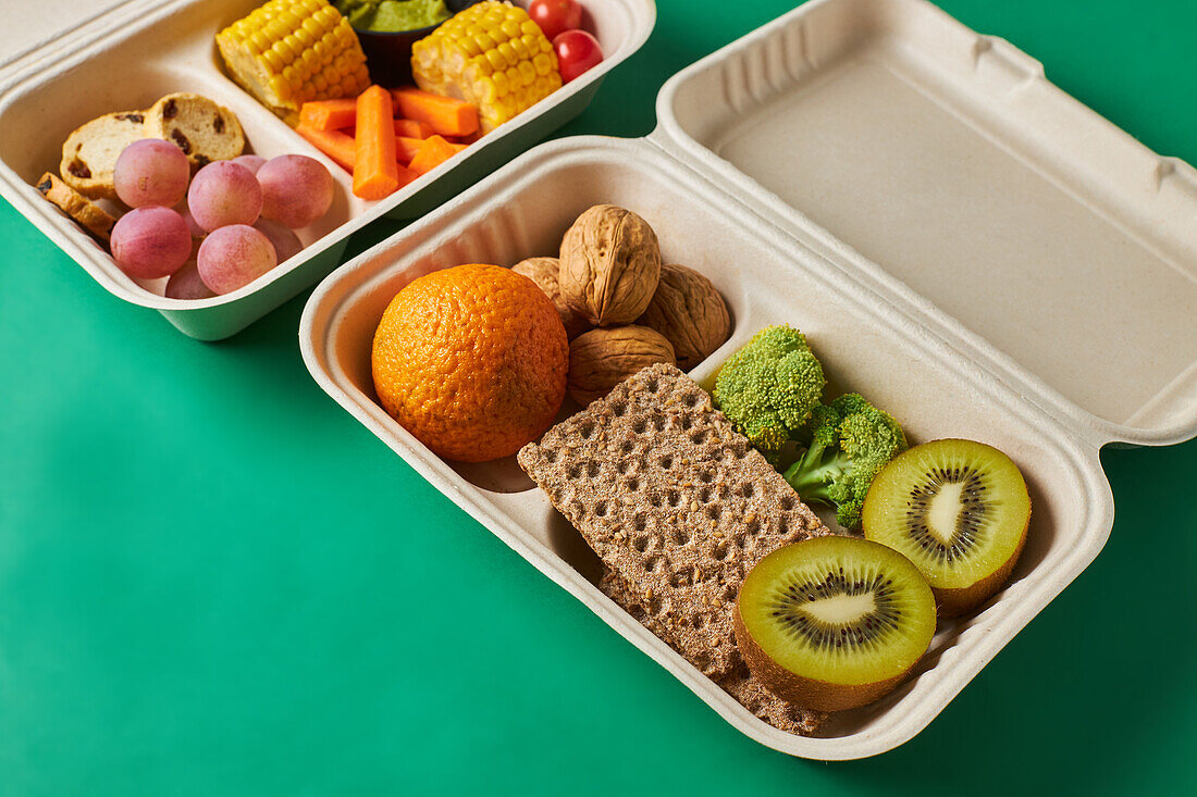 From above of lunch boxes with healthy food including crackers carrot sticks grapes cherry tomatoes with kiwi broccoli walnut and tangerine on green background