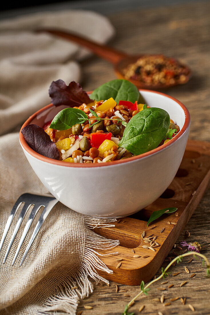 Ceramic bowl filled with healthy salad of beans nuts and vegetables supported by wooden board on table with fork