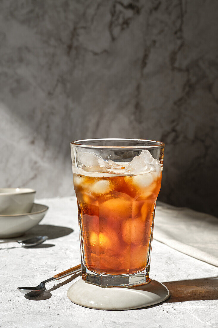 Glass of refreshing cold tea with ice cubes served on glass on table with spoon in sunlight