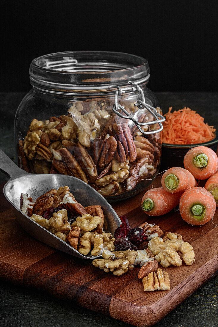 Glass jar with walnuts and dried fruits on a dark rustic background next to carrots, ingredients to prepare a carrot cake