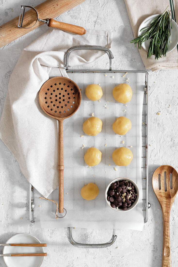 Top view of sweet cookie dough in shape of balls placed on baking paper on table with chocolate chips and rosemary branches before baking