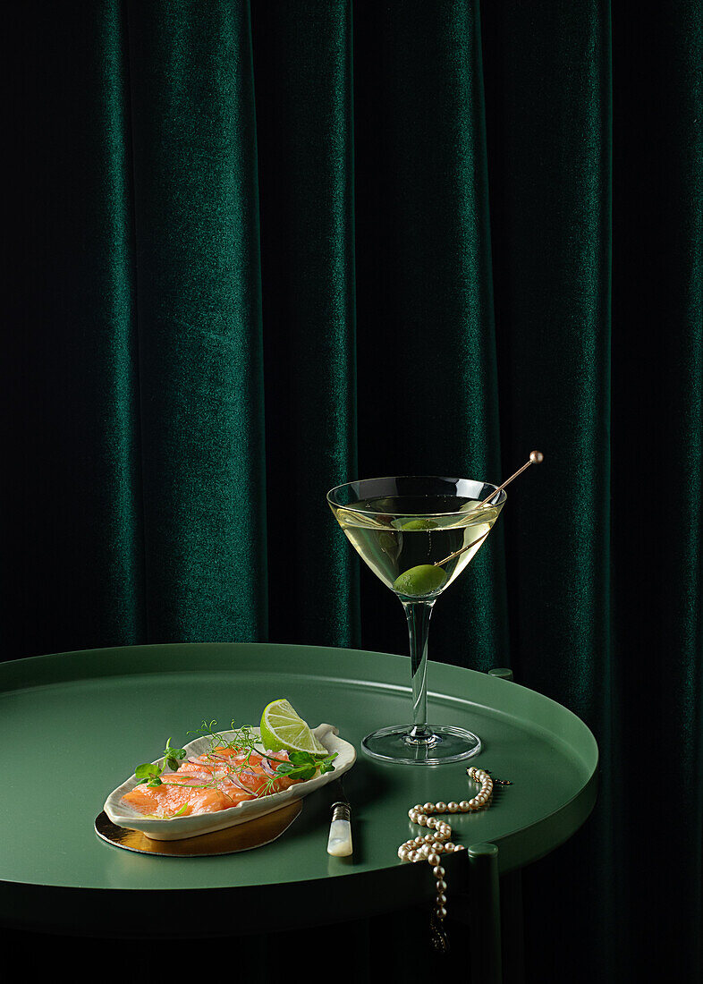 From above of appetizing smoked salmon slices served on plate with lime and onion and placed on round table with glass of vermouth with olive near elegant pearl necklace