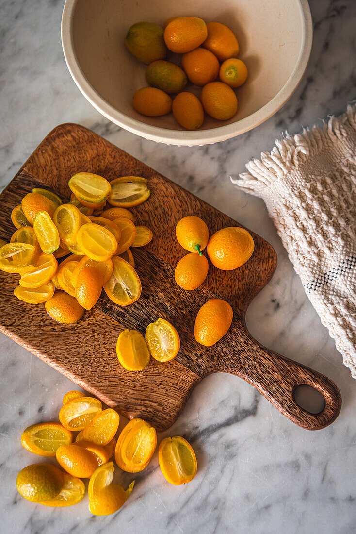 Draufsicht auf einen Stapel frischer, in Orangen geschnittener Kumquats auf einem hölzernen Schneidebrett auf einem Marmortisch mit Handtuch in der Küche
