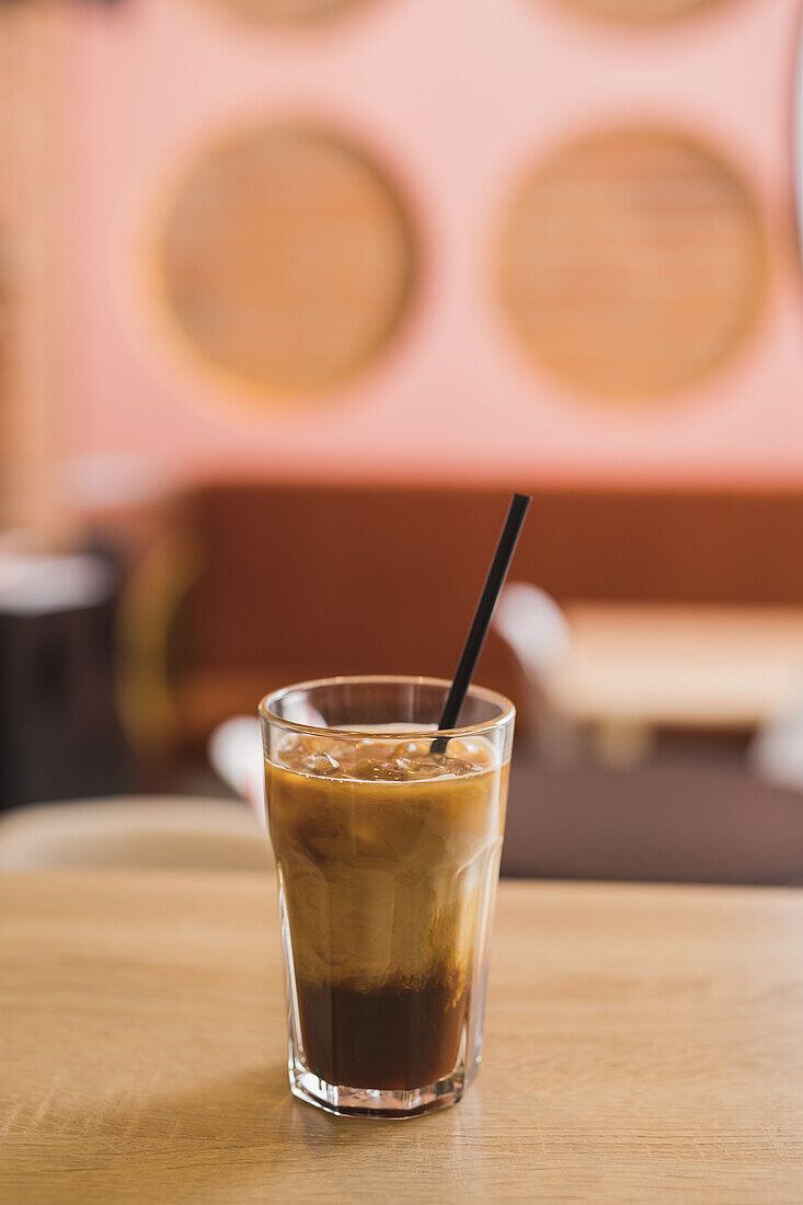 Hohes Glas mit schwarzem Eiskaffee mit sich langsam auflösender Frischmilch und mit Plastikstrohhalm auf dem Tisch in einem hellen Cafe