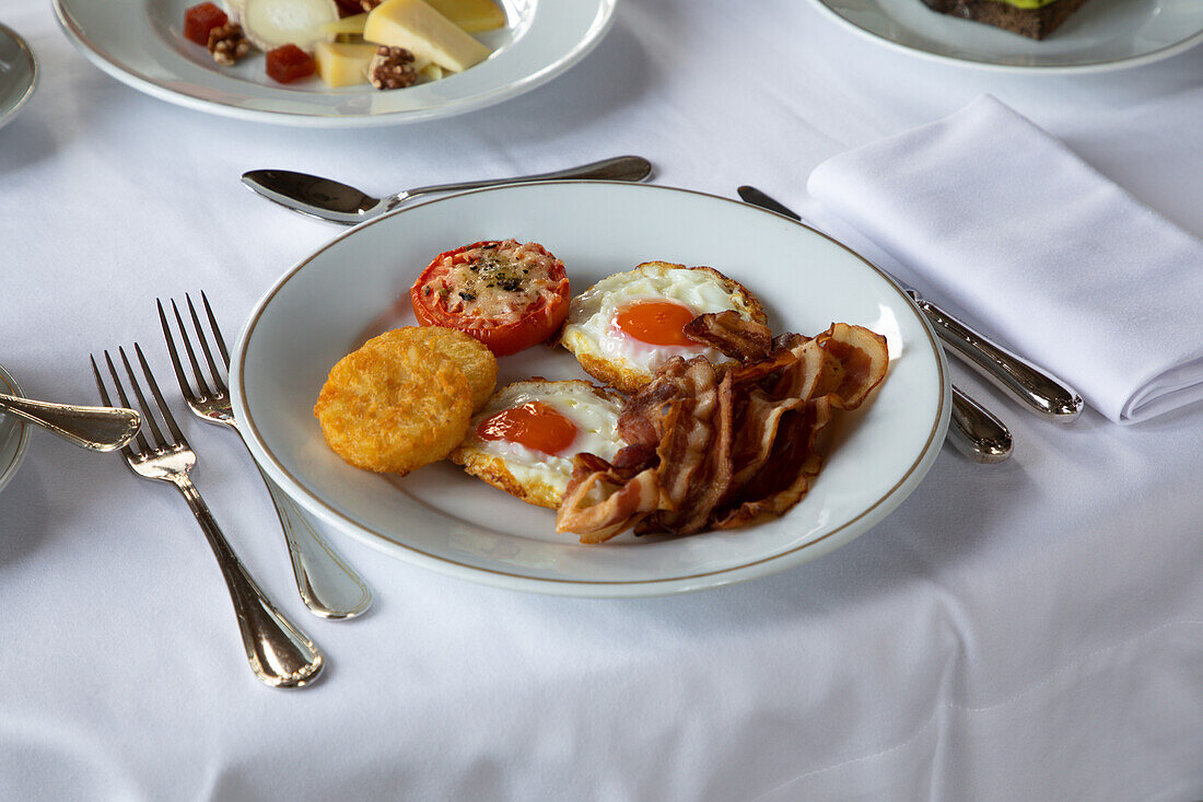 Appetitliche Spiegeleier mit Speckscheiben auf einem Teller mit gefüllter Tomate und knusprigem Mozzarella auf einem Tisch mit Besteck und einer Tasse Kaffee beim Frühstück