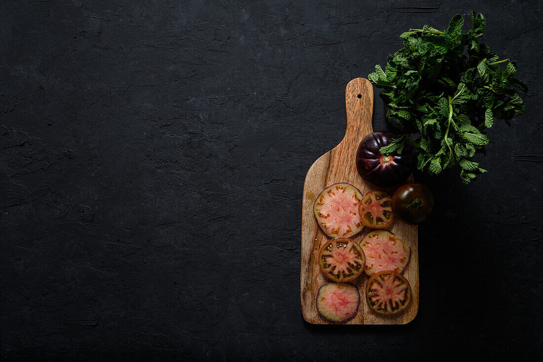 Top view of fresh ripe sliced black tomatoes and green mint stems on wooden cutting board on black background