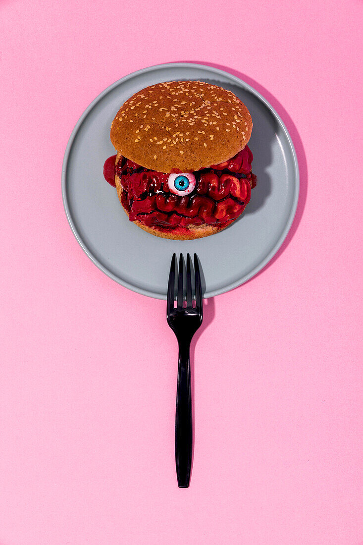 Draufsicht auf einen gruseligen Burger mit blutigem Gehirn und Auge, serviert auf einem grauen Teller mit Gabel vor rosa Hintergrund