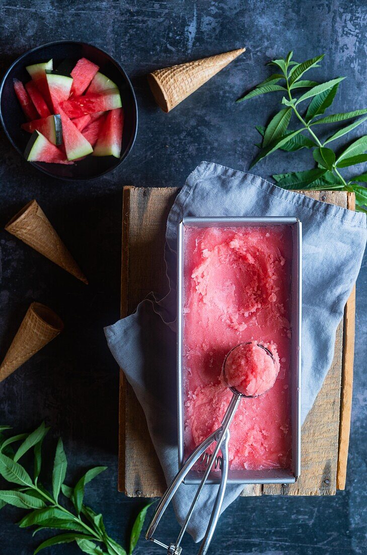 Draufsicht auf einen Behälter mit süßem Wassermelonensorbet und einem Löffel, der auf einer Serviette und einem Brett in der Nähe von Minzzweigen und Kornetts auf einem grauen Tisch steht