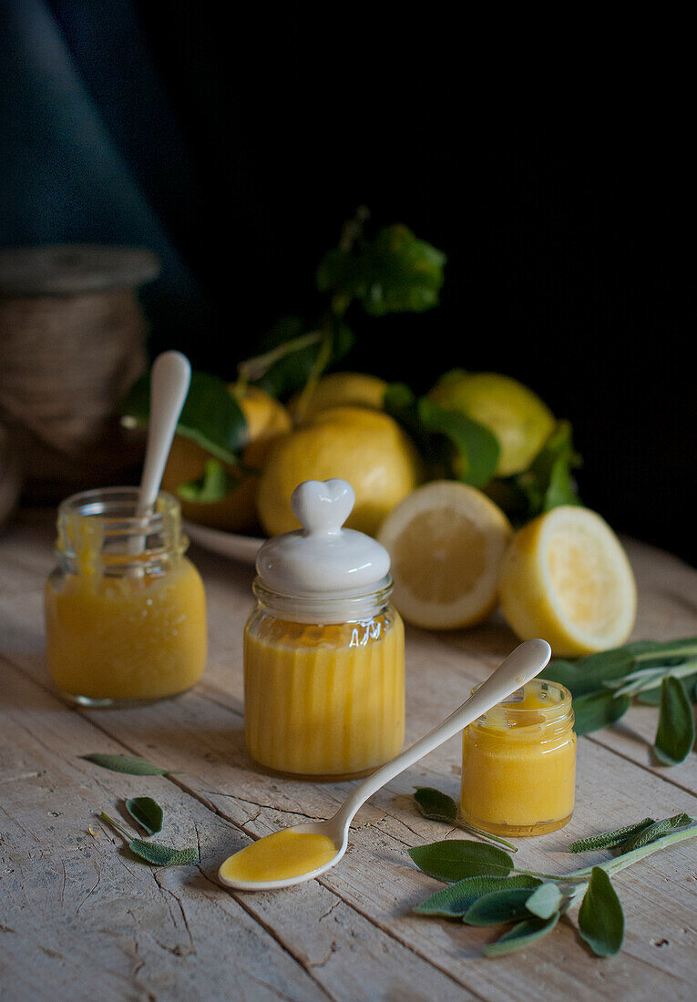 Small jars with yellow homemade lemon curd placed on wooden table with spoons and green leaves in kitchen at home