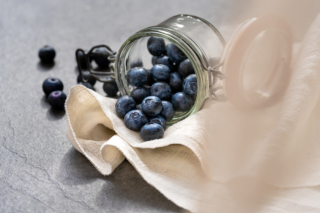 Pile of fresh ripe blueberries scattered from glass jar on napkin on gray table in studio