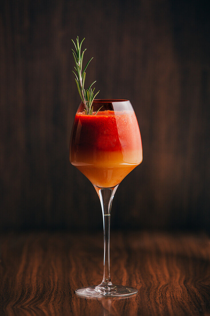 Close up view of red and orange cocktail with rosemary placed on wooden surface
