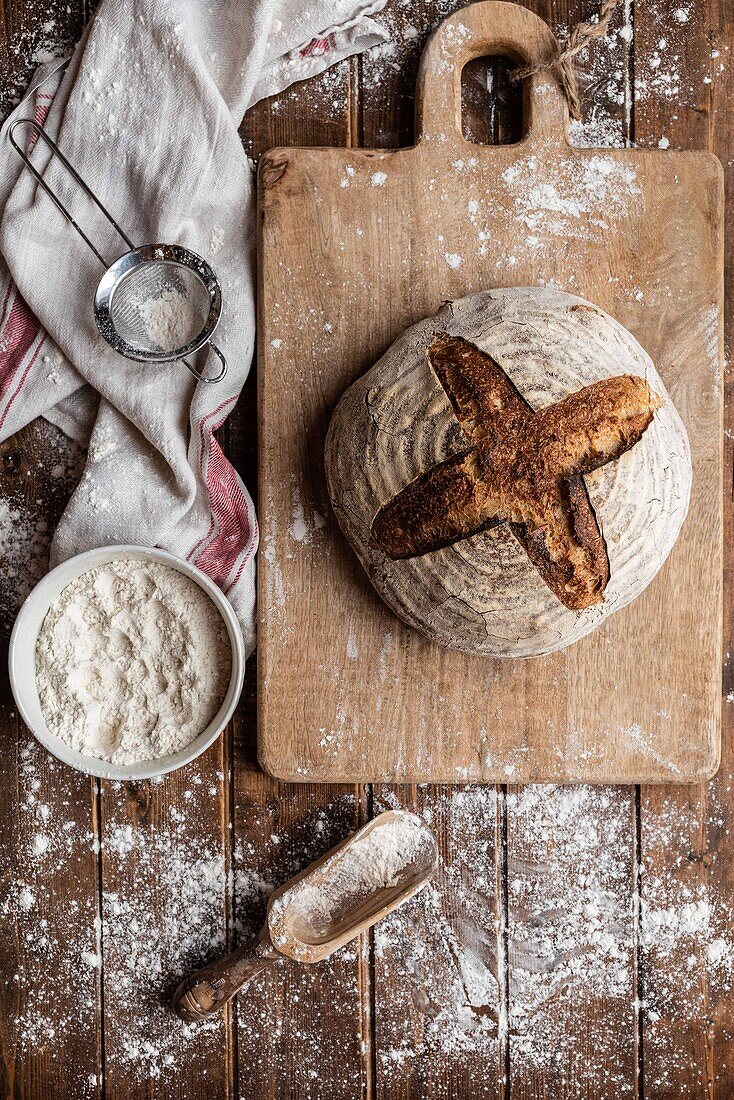 Draufsicht auf einen unordentlichen rustikalen Holztisch aus Weizenmehl und ein Brett mit frisch gebackenem Sauerteigbrotlaib