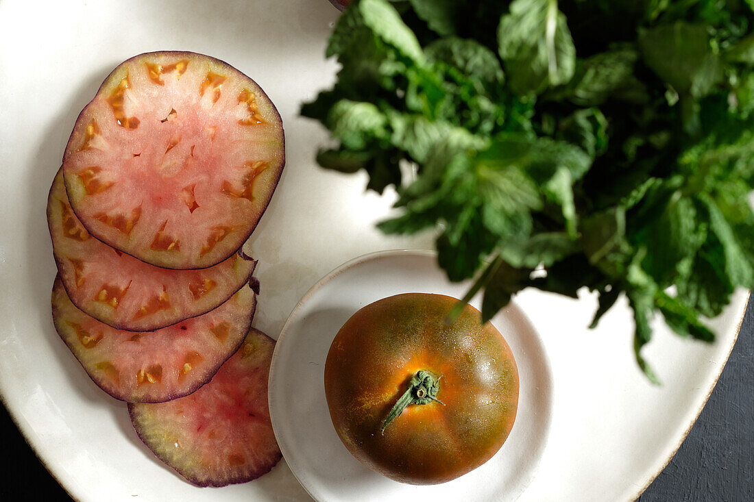 Draufsicht auf frische ganze und in Scheiben geschnittene schwarze Tomaten auf dem Tisch und grüne Minze bei der Zubereitung einer gesunden Mahlzeit