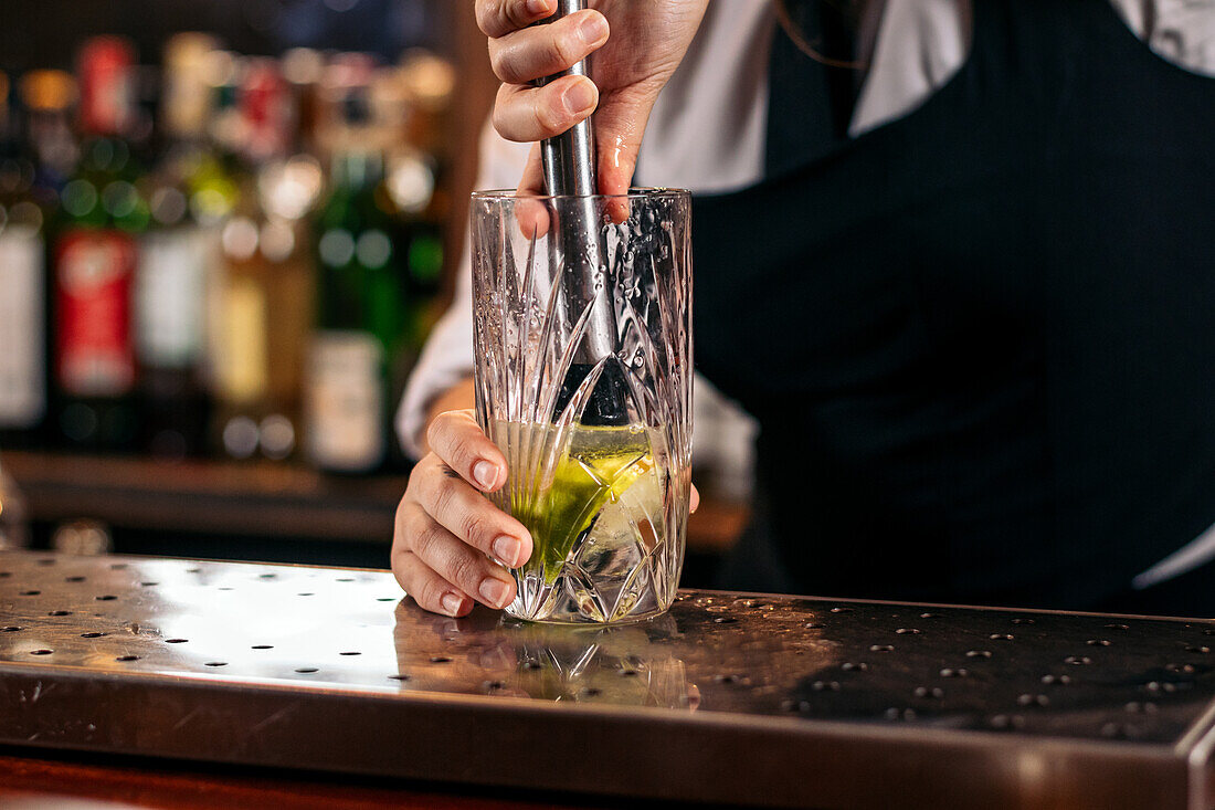 Crop unrecognizable barkeeper with muddler mixing refreshing cocktail in glass with ice and lime while working in pub