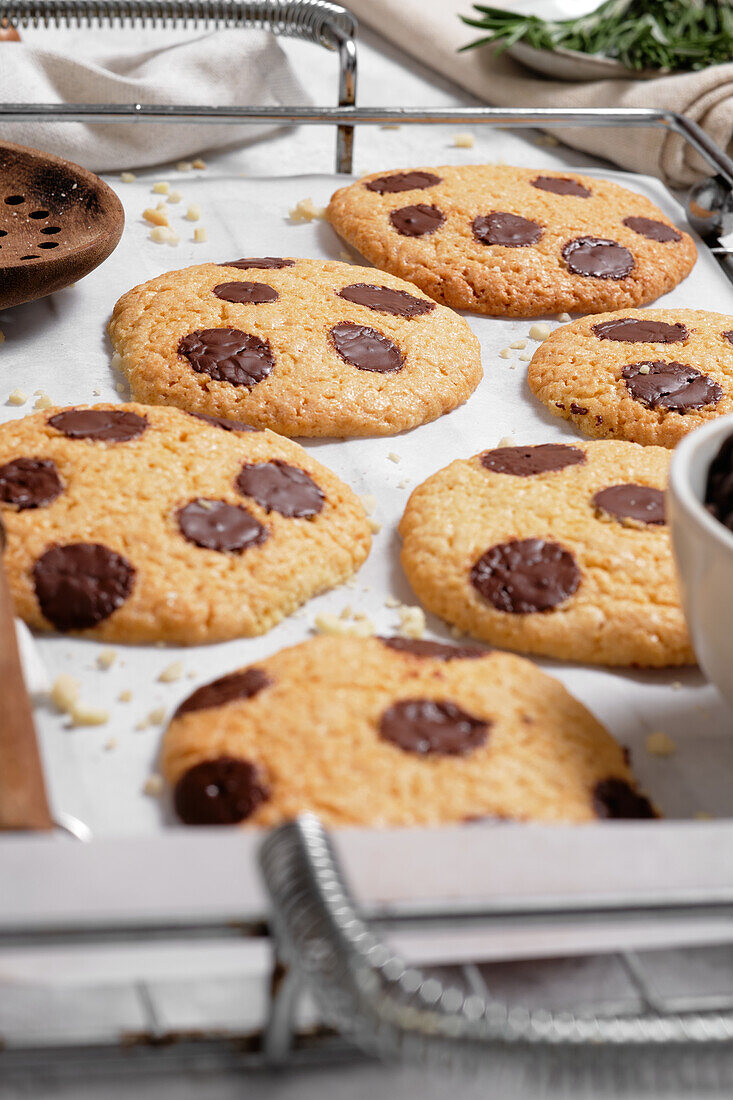 Blick von oben auf frisch gebackene süße Kekse mit Schokoladensplittern auf einem Metallgitter auf einem Tisch mit verschiedenen Küchengeräten und grünen Rosmarinzweigen